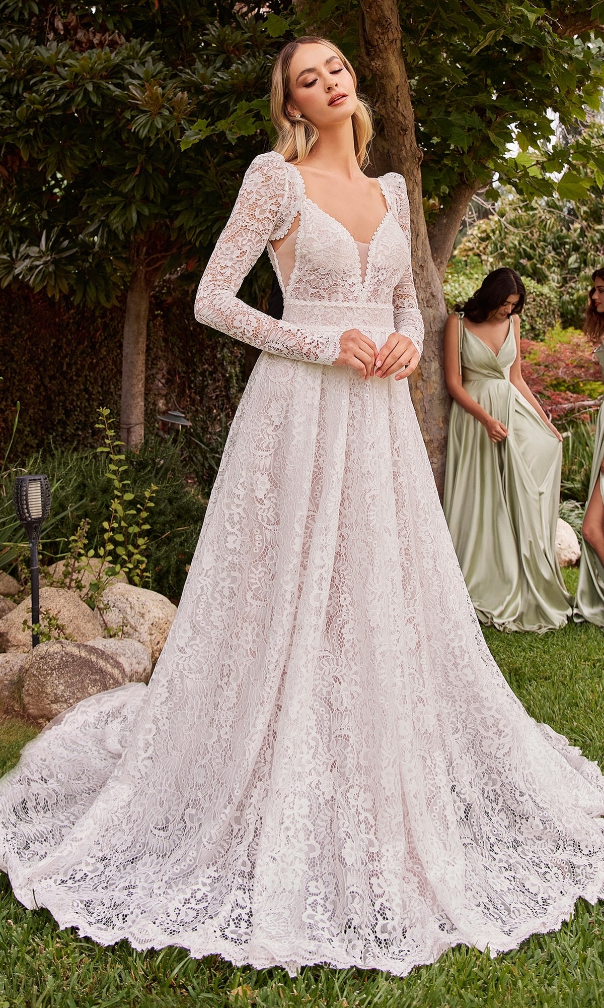 A woman stands outdoors in the Ladivine A-Line Lace Wedding Dress CD862W with a train; two women in sage green dresses are in the background.